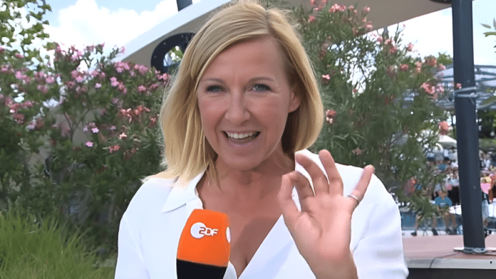 Andrea Kiewel, die Moderatorin des ZDF-Fernsehgartens, lächelt in die Kamera. Sie trägt ein weißes Oberteil und hält ein ZDF-Mikrofon in der Hand. Im Hintergrund sind blühende rosa Blumen zu sehen. Kiewel winkt fröhlich mit ihrer freien Hand in Richtung Kamera. Die Szene spielt sich im Freien ab, vermutlich am Set des Fernsehgartens.