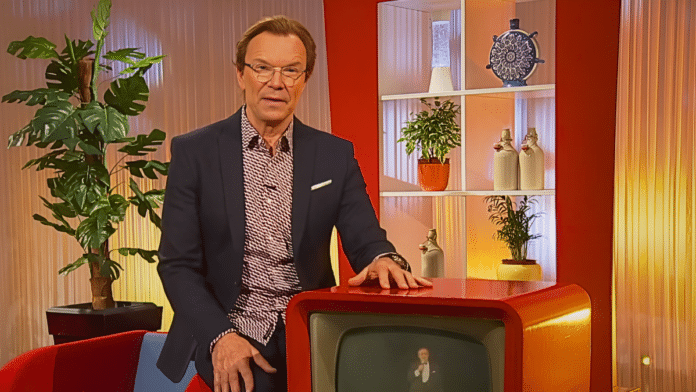 Wolfgang Lippert steht in einem TV-Studio, gekleidet in dunklem Blazer und gemustertem Hemd. Er lehnt an einem roten Retro-Fernseher. Im Hintergrund sieht man ein gemütliches Set-Design mit Pflanzen, Regalen und dekorativen Elementen. Die Atmosphäre ist warm und einladend, mit orange-roten Farbtönen und weicher Beleuchtung.